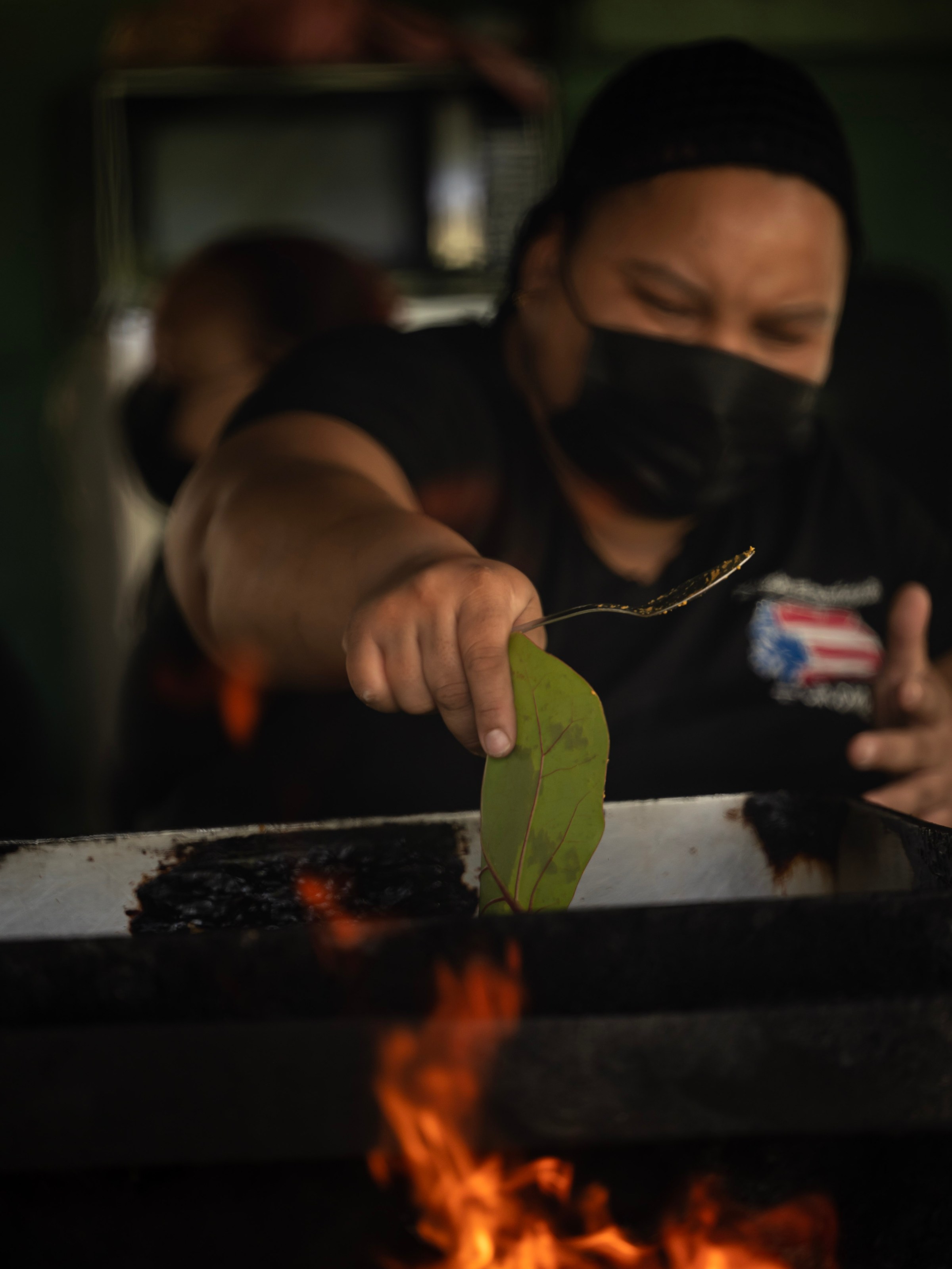 Person with mask places a green leaf over flames on a grill.