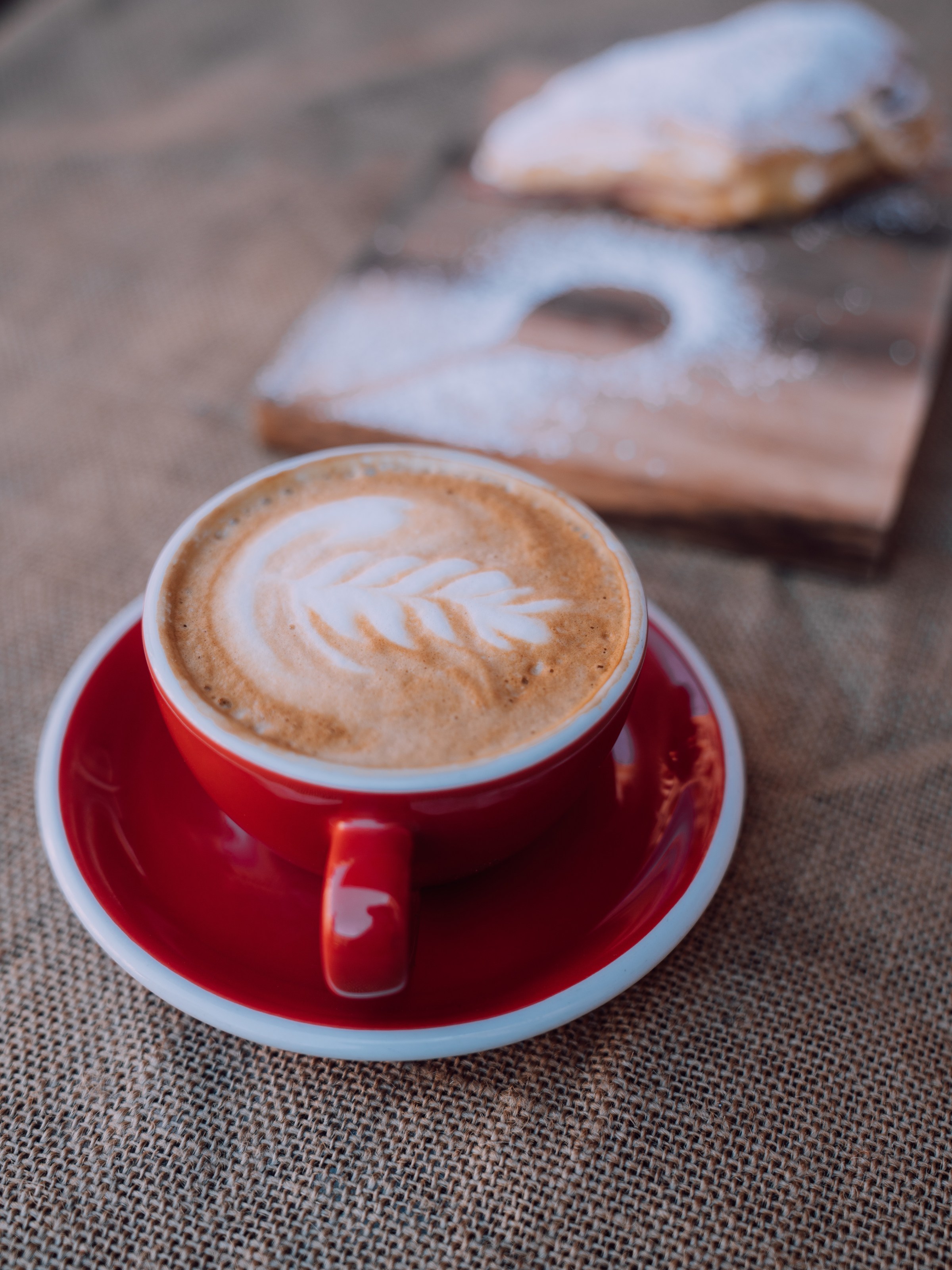 Latte in a red cup with latte art on burlap, pastry in background.