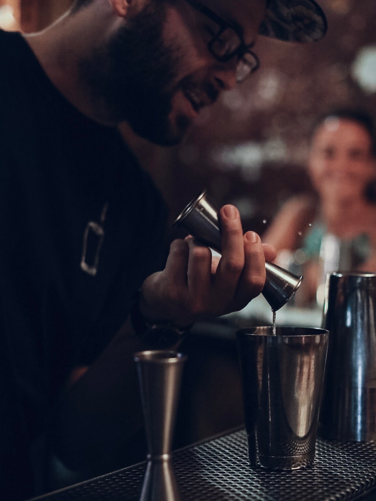 Bartender pouring liquid into a shaker with a jigger, smiling woman in the background.