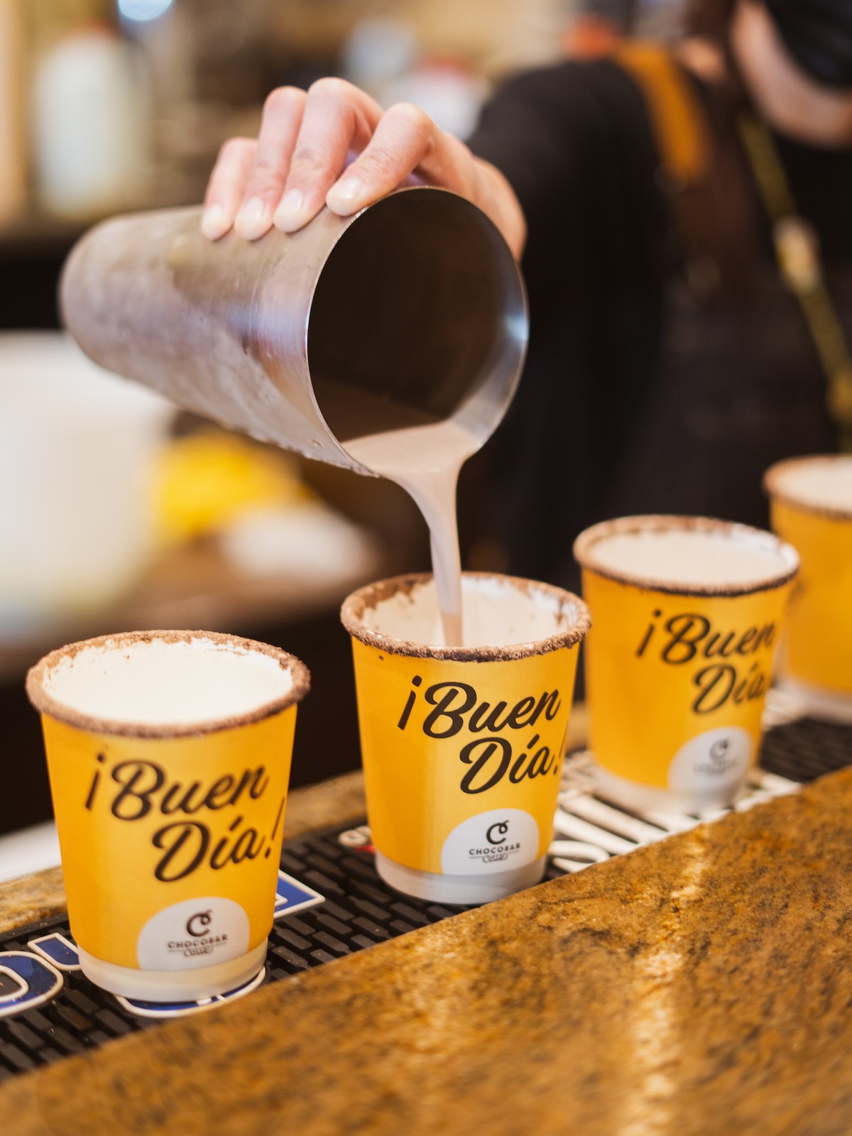 A hand pouring liquid into four yellow cups labeled '¡Buen Día!' on a countertop.