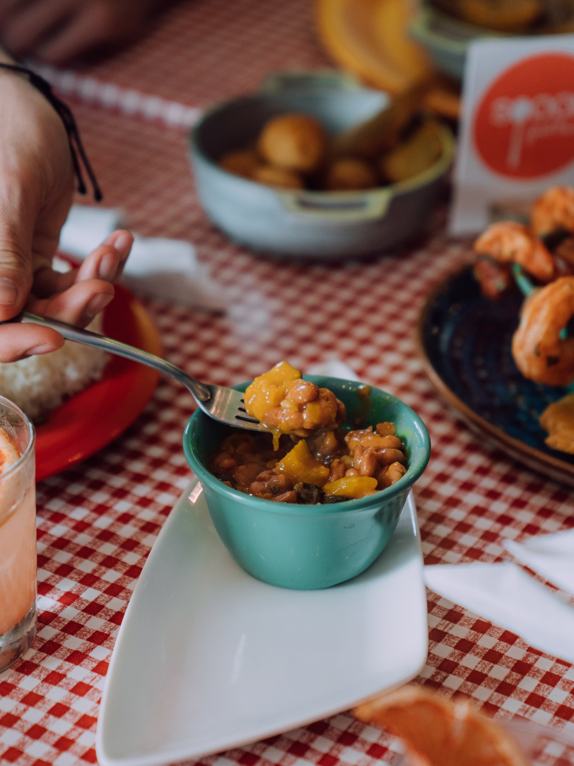 Hand holding fork over food on checked tablecloth near drink and assorted dishes.