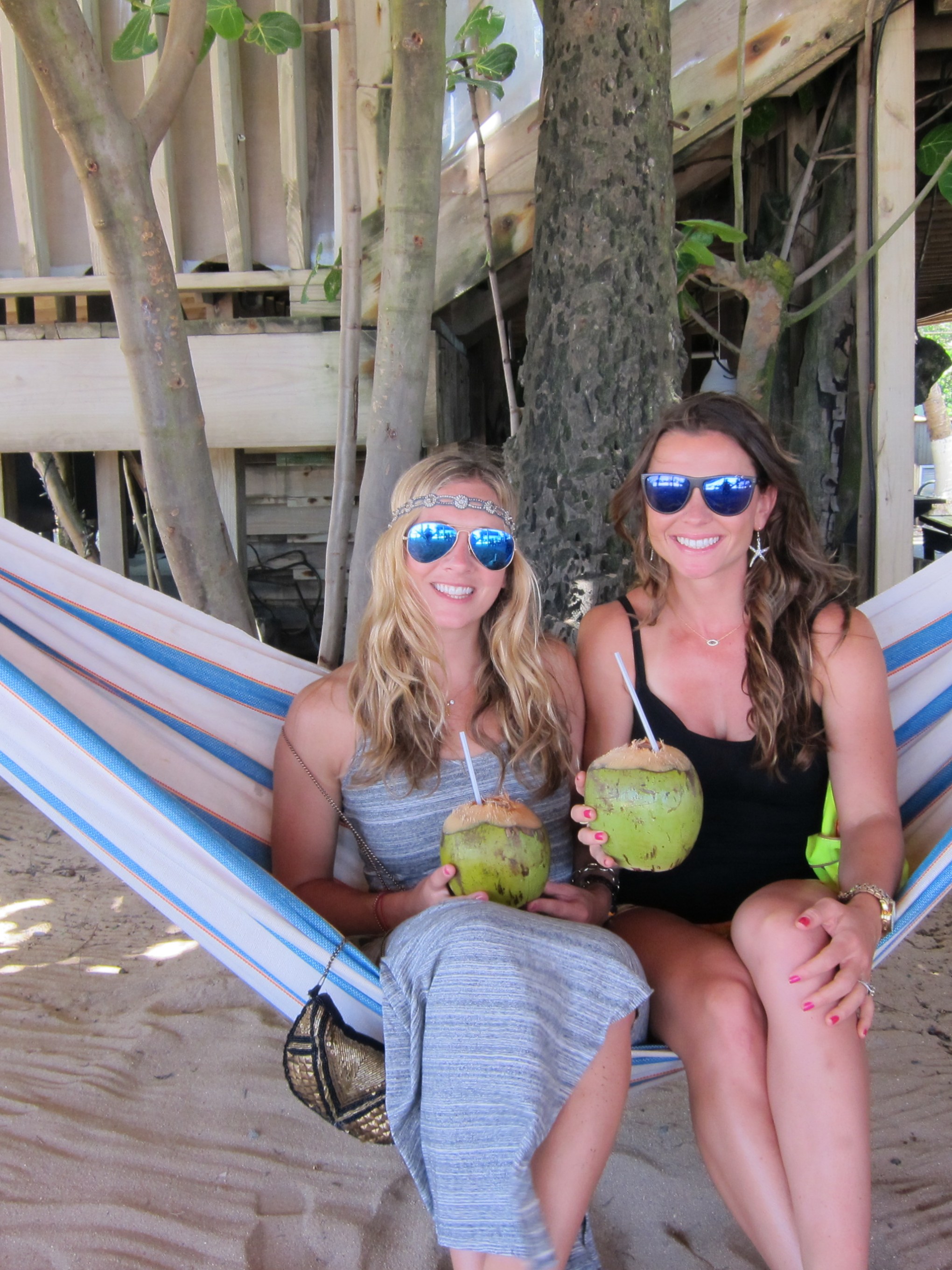 Two people in sunglasses sit on a hammock holding coconuts with straws on a sandy beach.