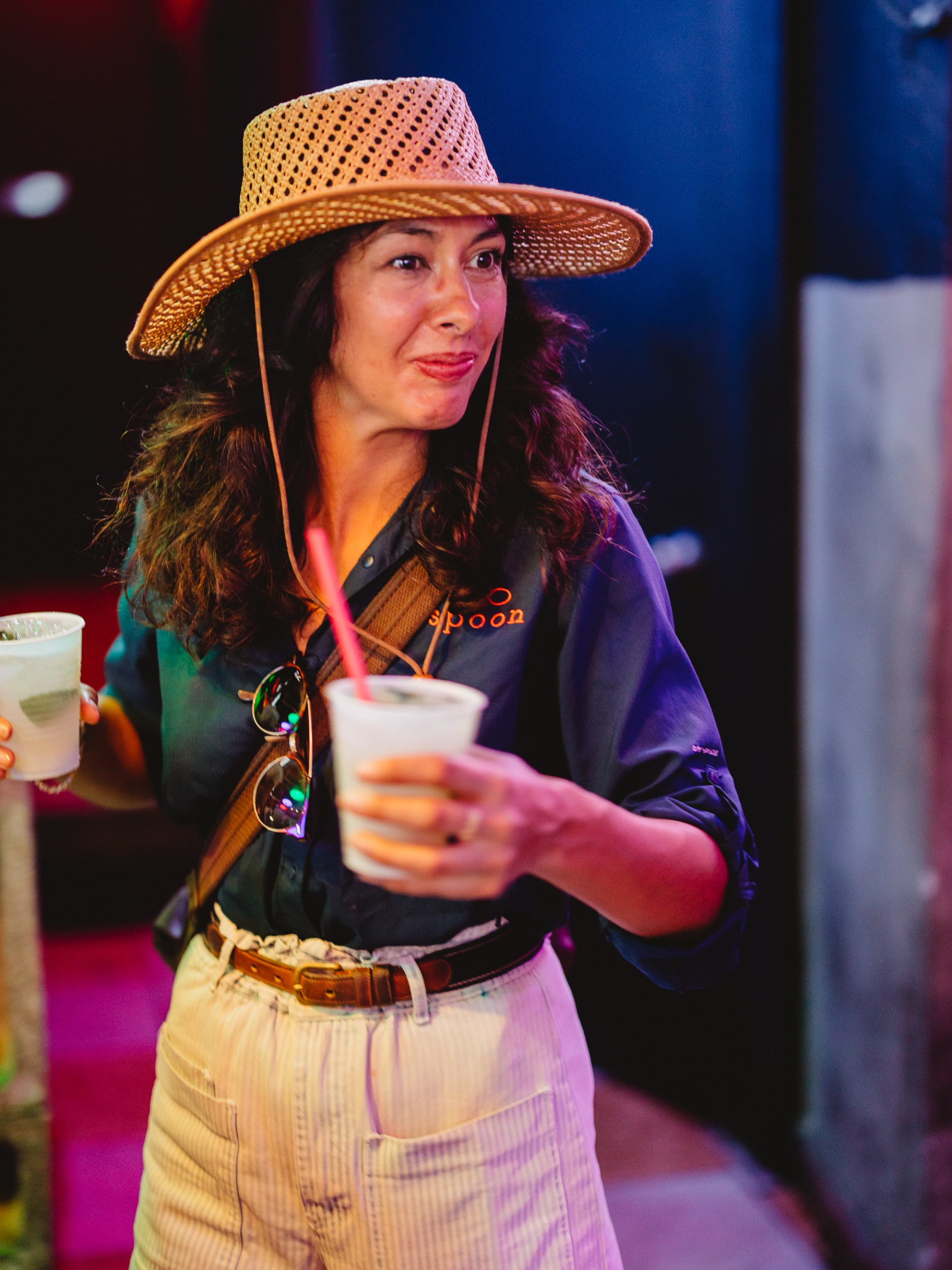 Woman in straw hat holding two drinks with striped shorts and sunglasses.