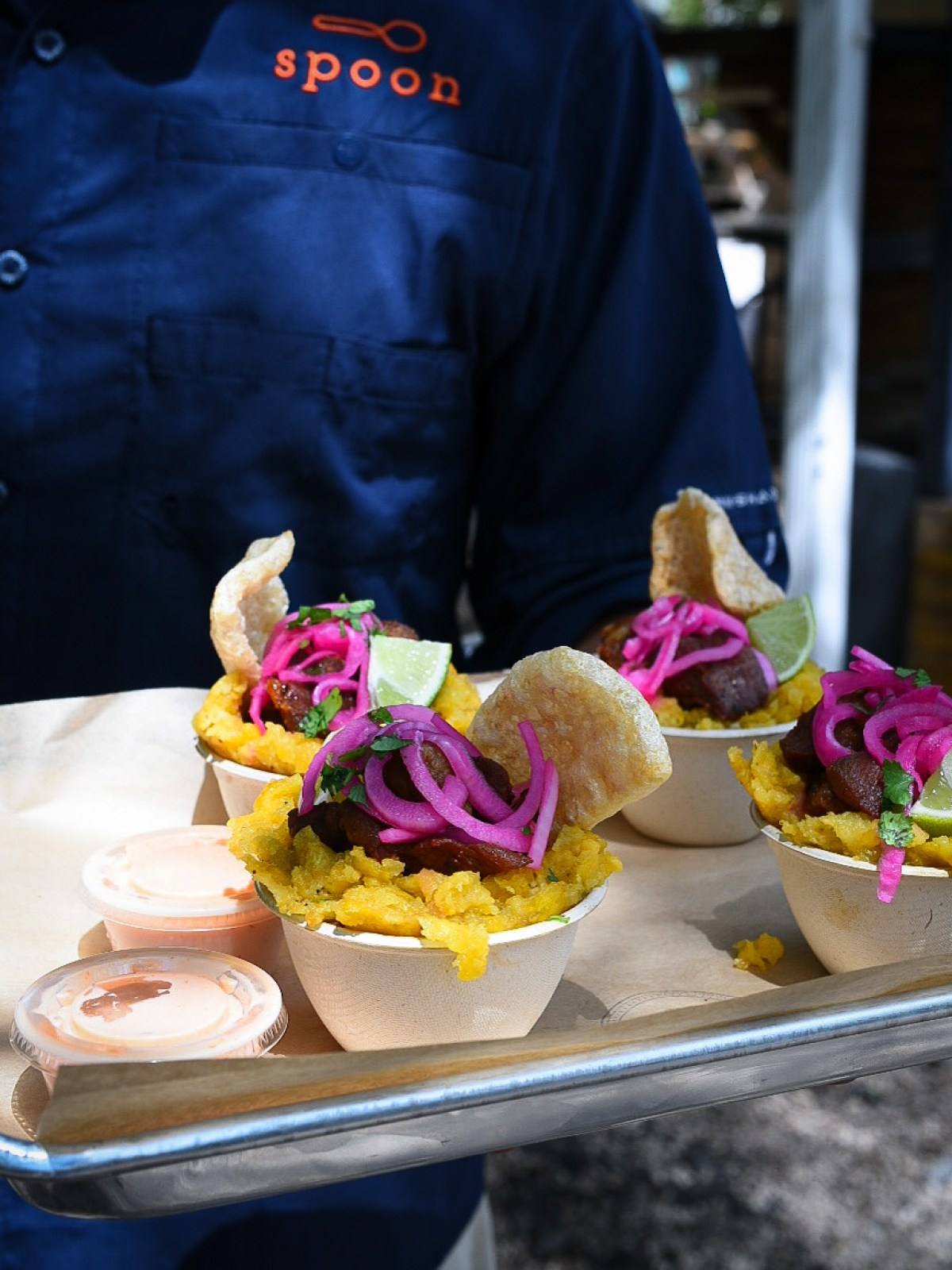 Chef in blue uniform holding a tray with bowls of colorful food topped with pink onions and lime wedges.