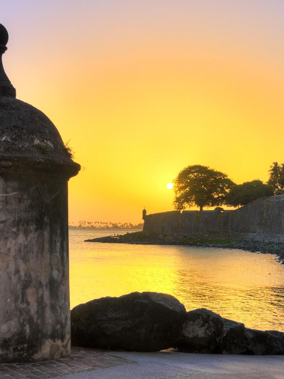 Sunrise at a coastal fort with stone walls and a turret, overlooking calm water.