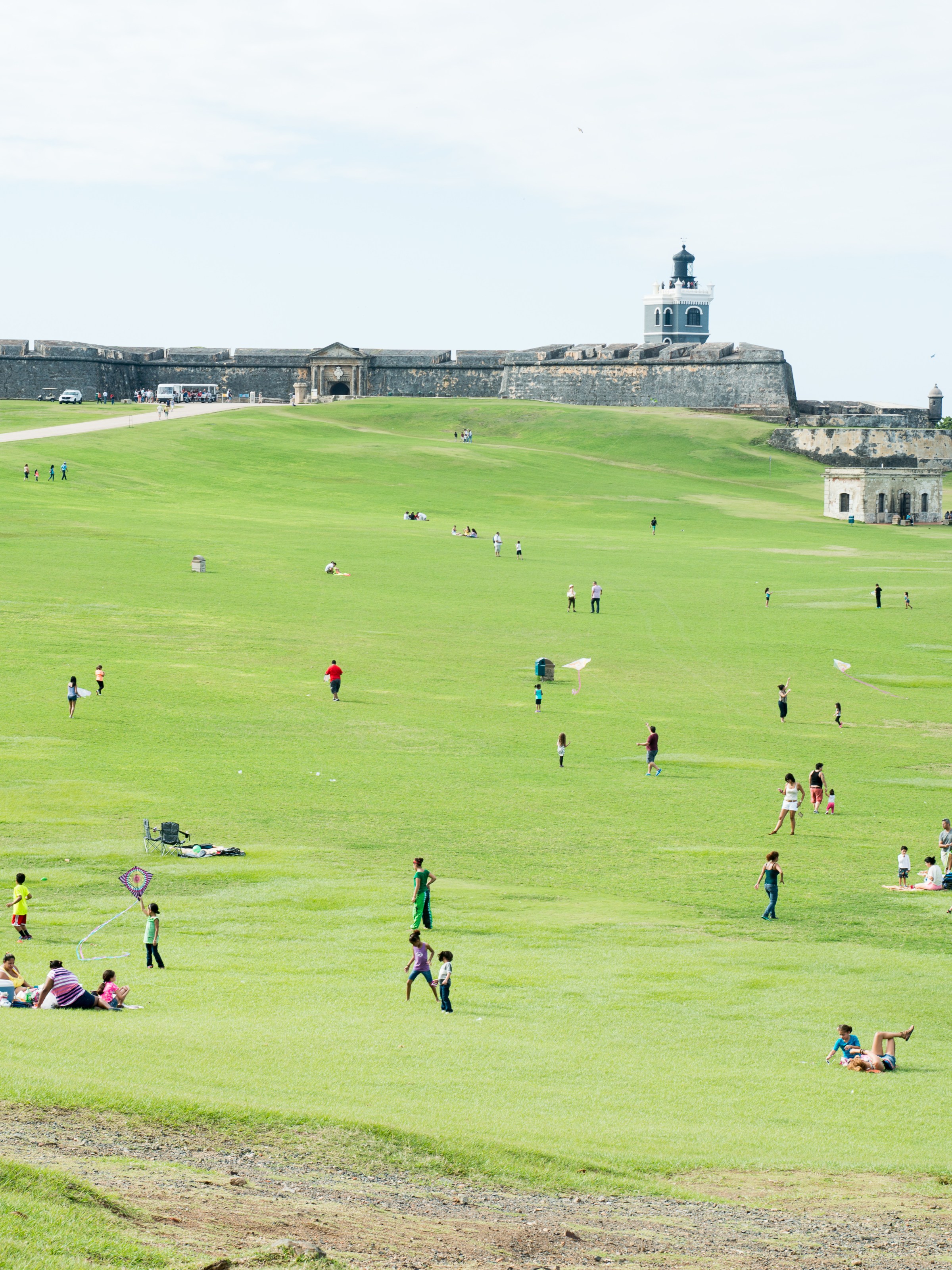 People enjoy a sunny day flying kites near an old fort on a grassy hillside.