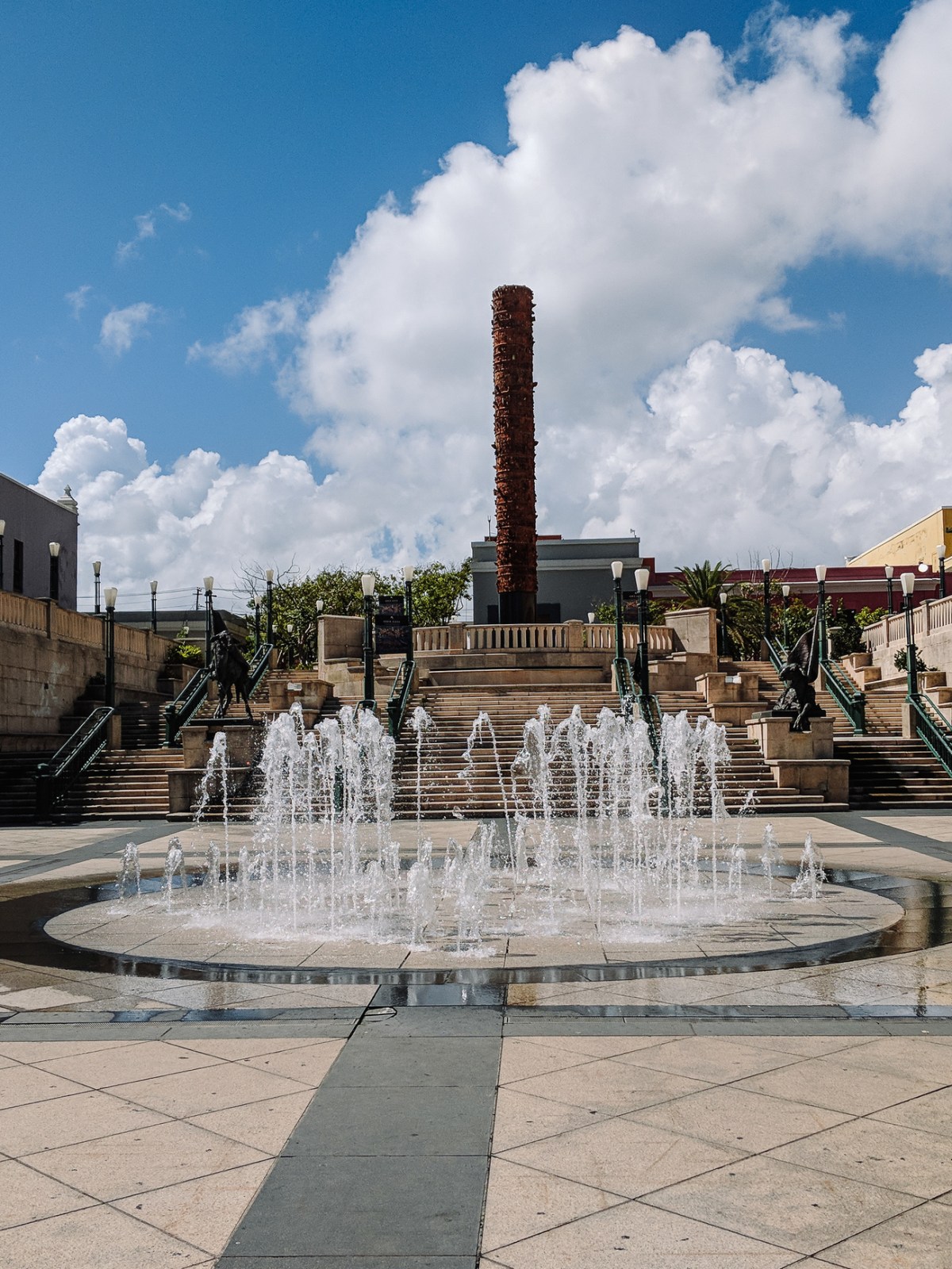 Fountain in a plaza with stairs and a tall, cylindrical sculpture under a blue sky with clouds.