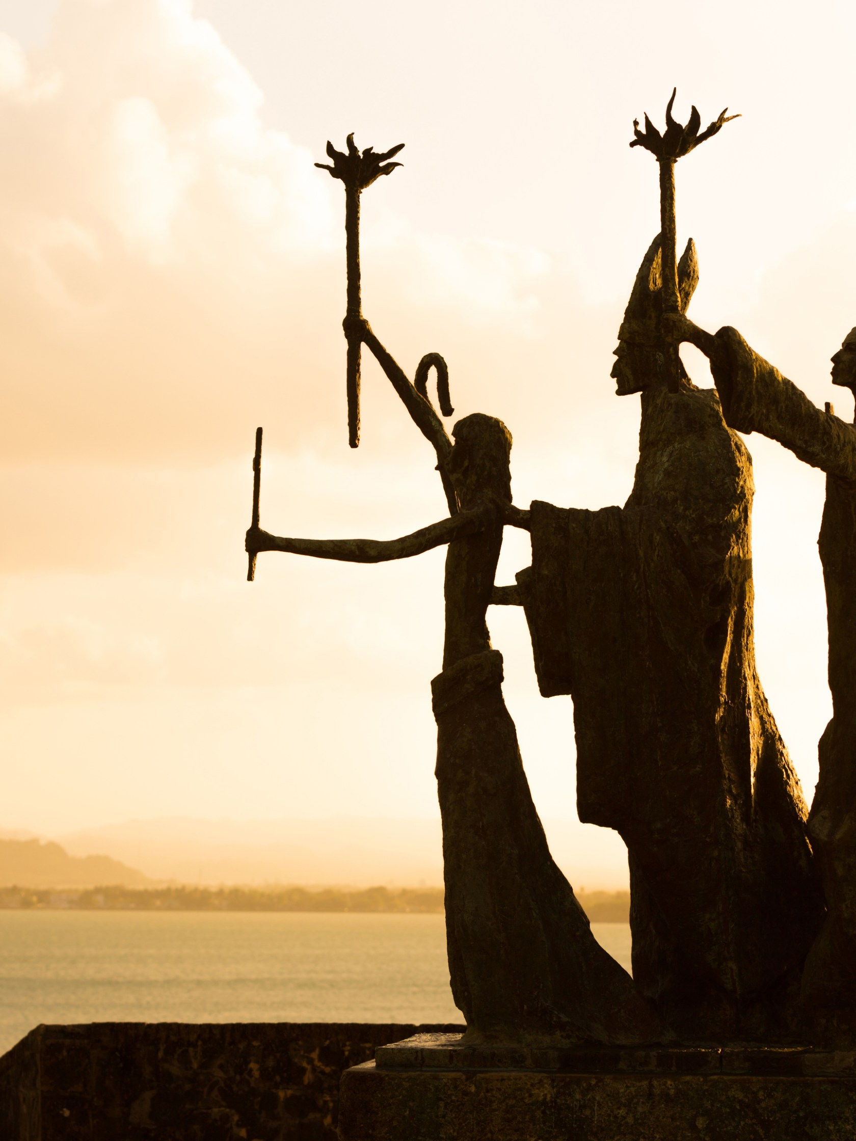 Silhouetted statues holding torches with ocean and sunset in background.