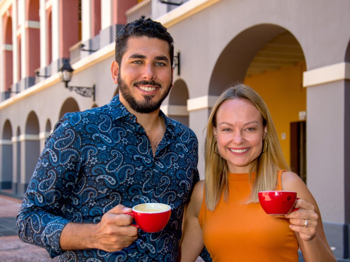 PaulinaSalachGustavoAntonetti (1) Two people smiling, holding red coffee cups in front of a colorful building.