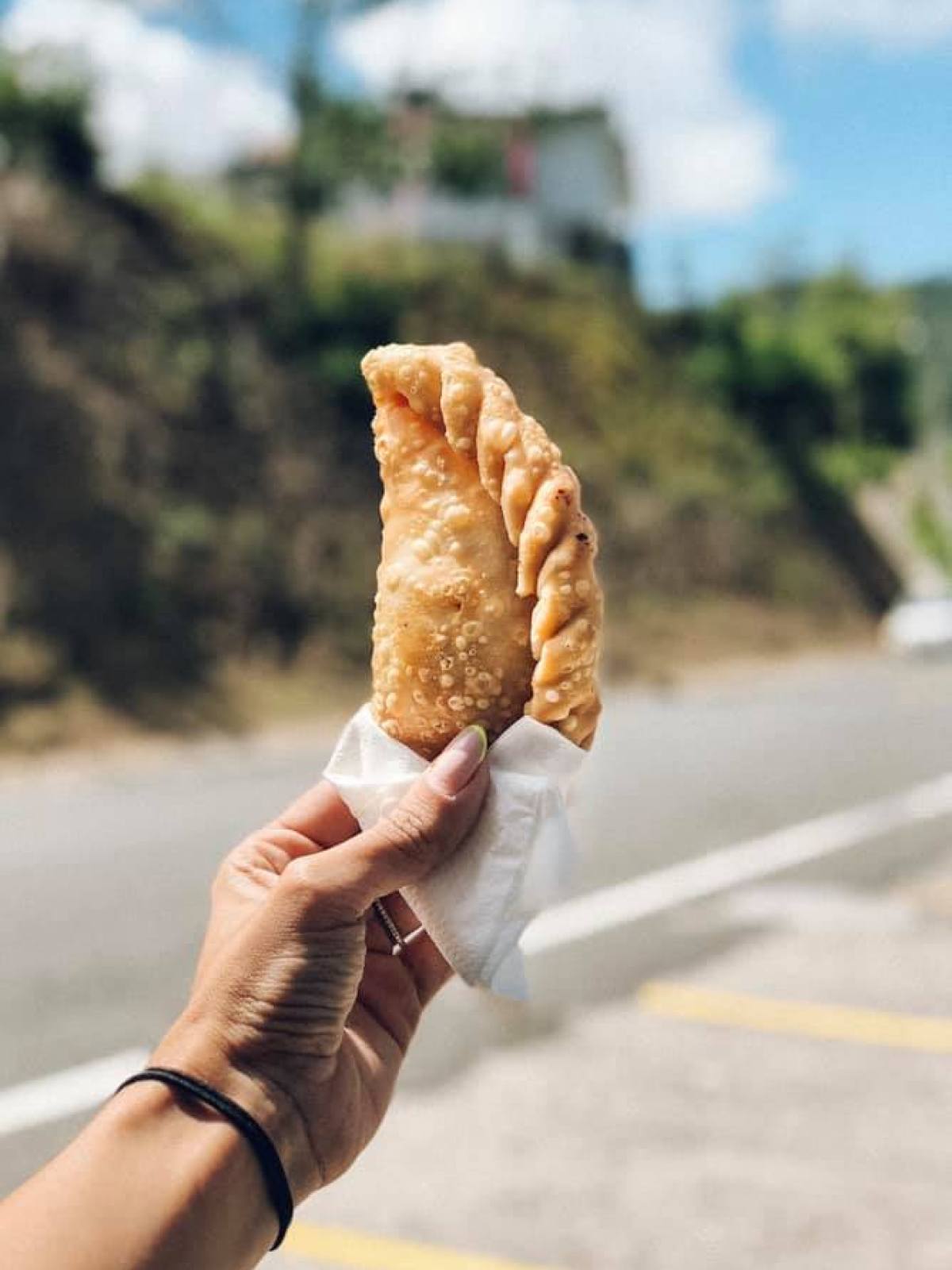 Hand holding a fried pastry with a napkin, outdoors with a road in the background.