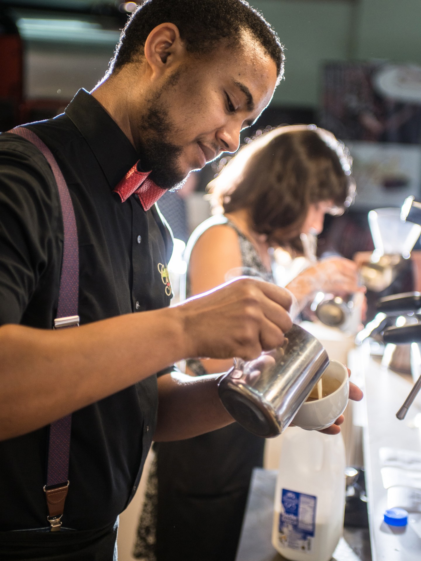 Barista pouring milk into a cup with another person preparing coffee in the background.