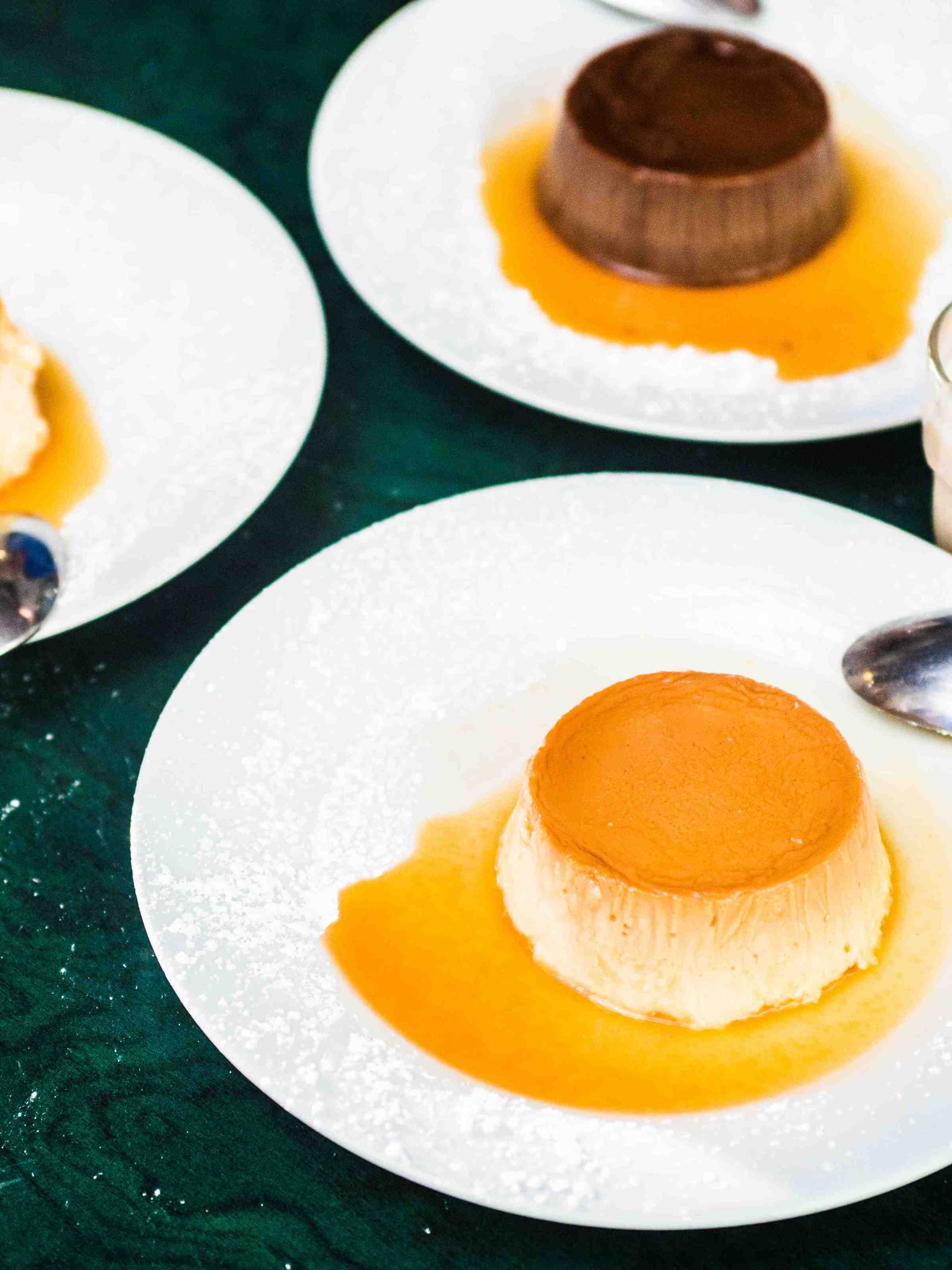 Three caramel flans on plates with spoons, one with a glass of milk.