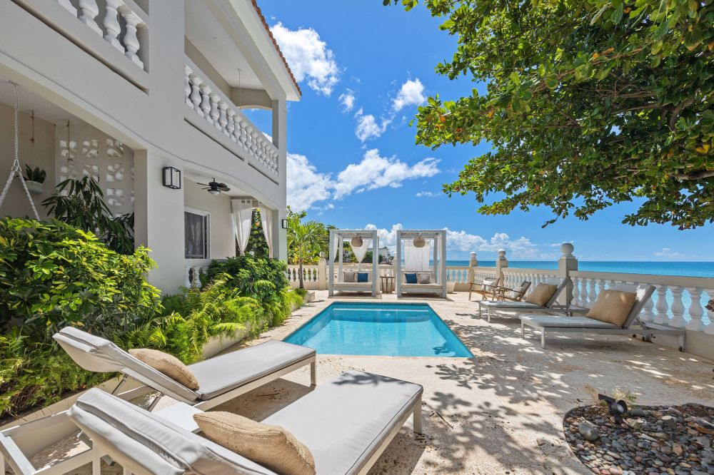 Beachfront patio with pool, lounge chairs, and ocean view, under a partly cloudy sky.