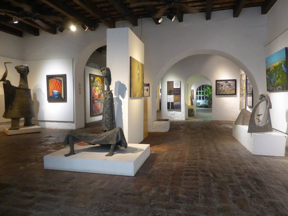 Art gallery with sculptures, paintings, and arched doorways on a terracotta-tiled floor.