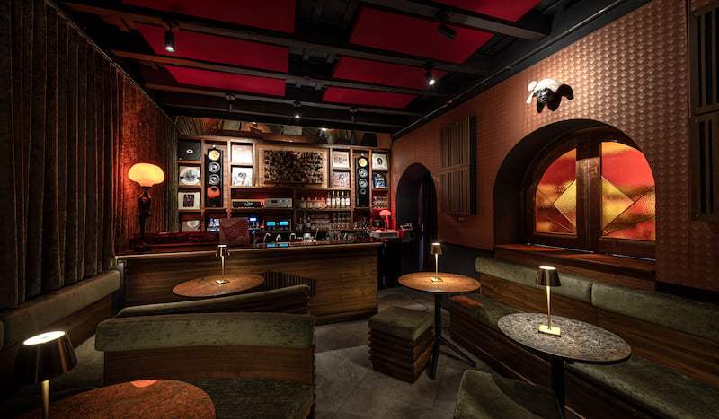 Cozy, dimly-lit bar with wooden decor, green seating, and small round tables, red ceiling accents.
