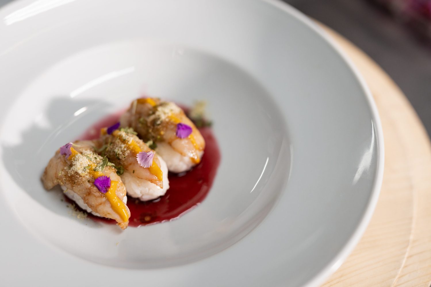 Three pieces of sushi on a white plate with red sauce and garnished with flowers.